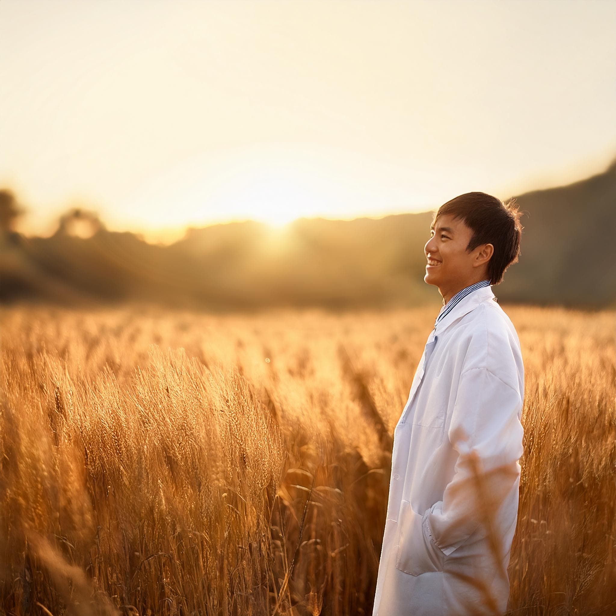 優しそうな笑顔のやせ型の日本人男性、白衣を羽織り金色に光り輝く畑を見つめている様子だが、逆光で顔は良く見えない